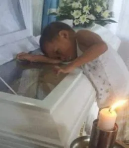 P1. Child silently approaches mother's coffin, presses head against it during service